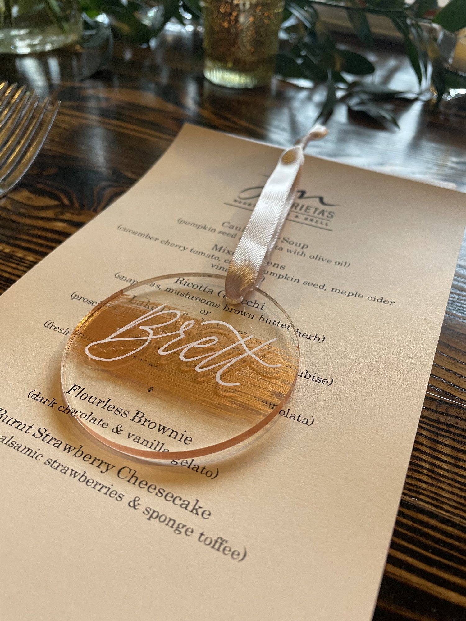 Hand-lettered acrylic place tag reading Brett, resting on a printed dinner menu